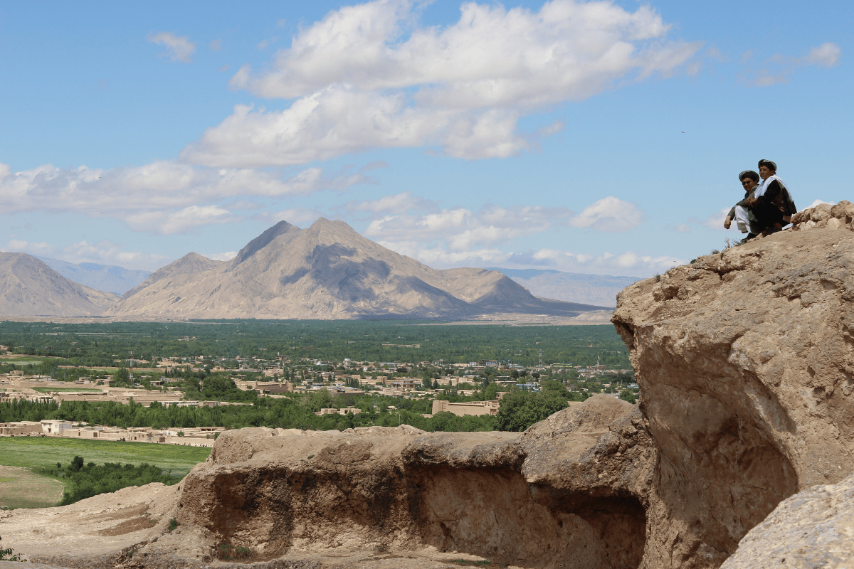 Two men sitting on a cliff in the foreground, with green fields and mountainous landscape in the background.