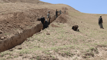 Dry grass landscape and people standing in line, digging a long, narrow ditch into the ground.
