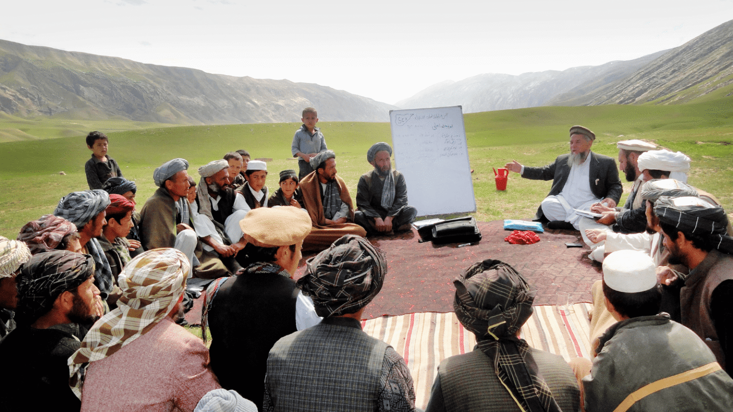 A large group of people are sitting in a circle on a carpet outside. One man, sitting next to a whiteboard, is pointing with one hand and speaking, while the rest of the group is paying attention. In the background, there is a green grass landscape and mountains.