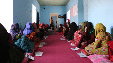 A group of women in colourful clothes, sitting on the floor in a room with blue walls.