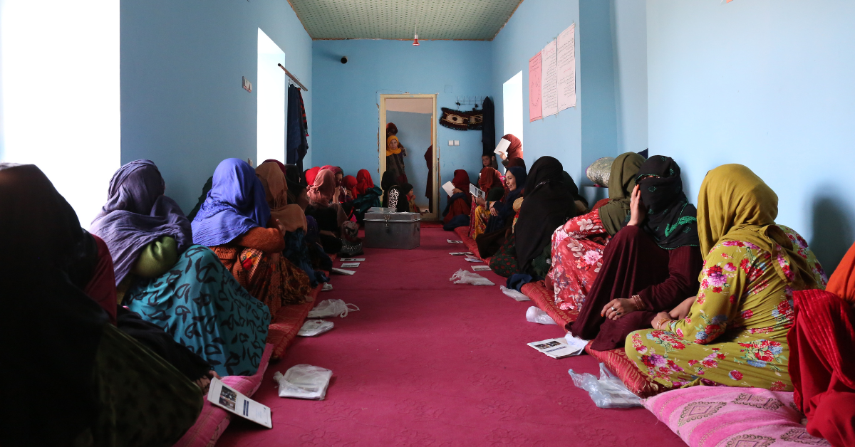 A group of women in colourful clothes, sitting on the floor in a room with blue walls.