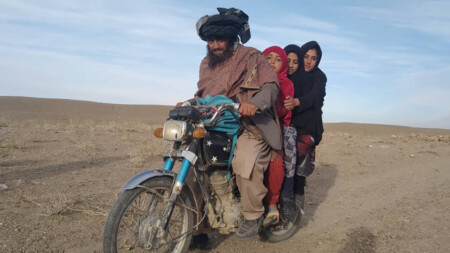 A man on a motorbike with three girls as passengers, in a dry Afghan landscape.