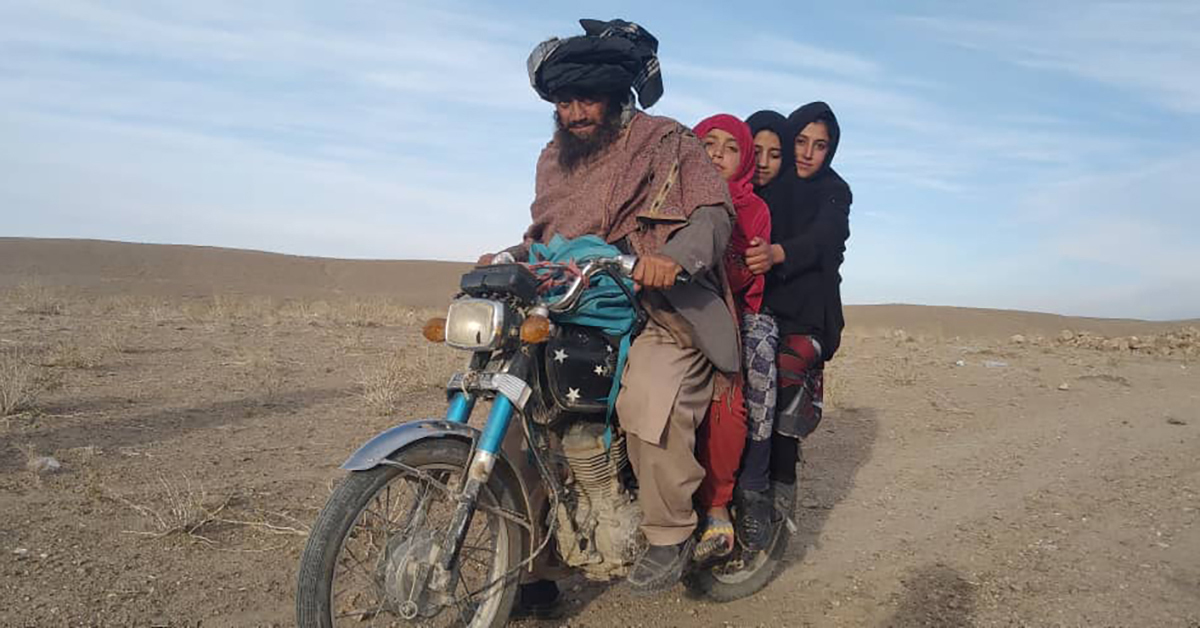 A man on a motorbike with three girls as passengers, in a dry Afghan landscape.