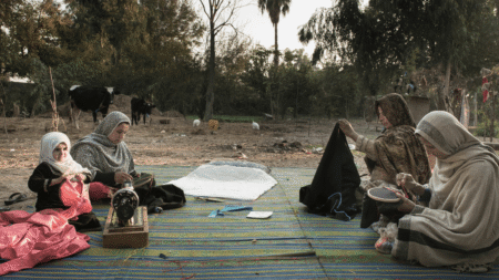 A group of women, sitting outside, on a carpet. They are sewing and embroidering. There is a sewing machine.