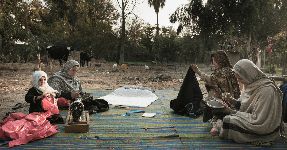 A group of women, sitting outside, on a carpet. They are sewing and embroidering. There is a sewing machine.