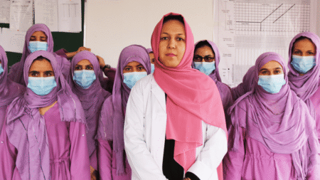 Hijabi women dressed in pink and purple clothes, wearing face masks and professional work clothes. At the front, in the middle, there is a woman in a white medical gown and pink hijab without a face mask.