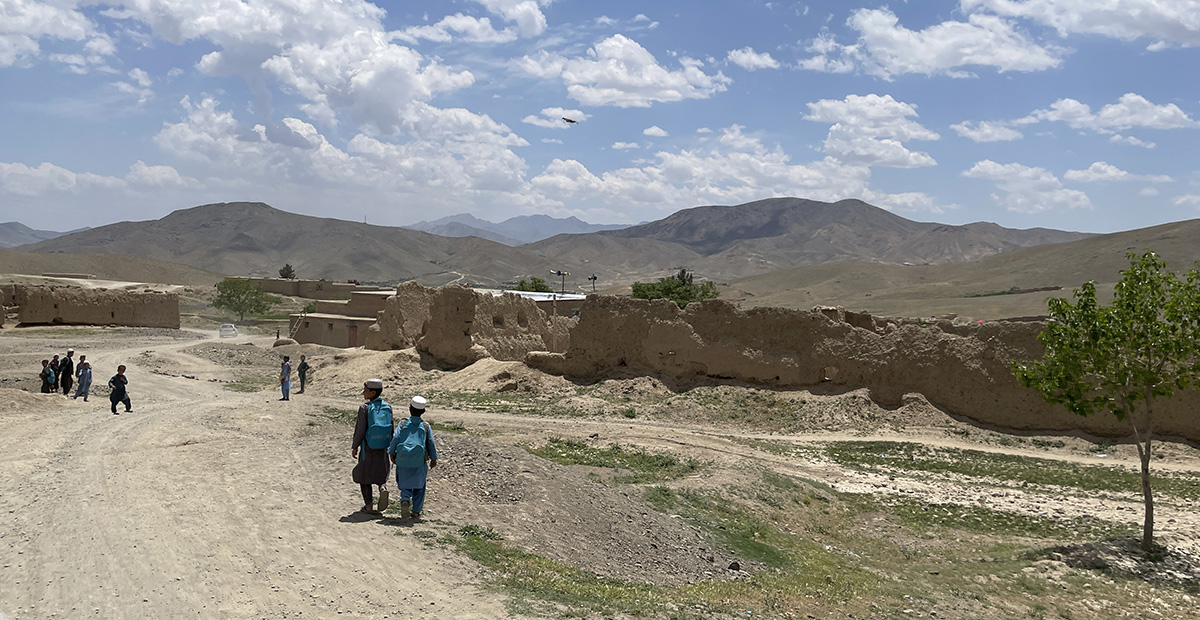 Schoolboys on a road in the Afghan countryside, with buildings close and mountains in the background.