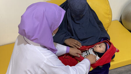 A woman with a purple shawl and a woman covered in a black shawl are caring for a baby wrapped in a red blanket.