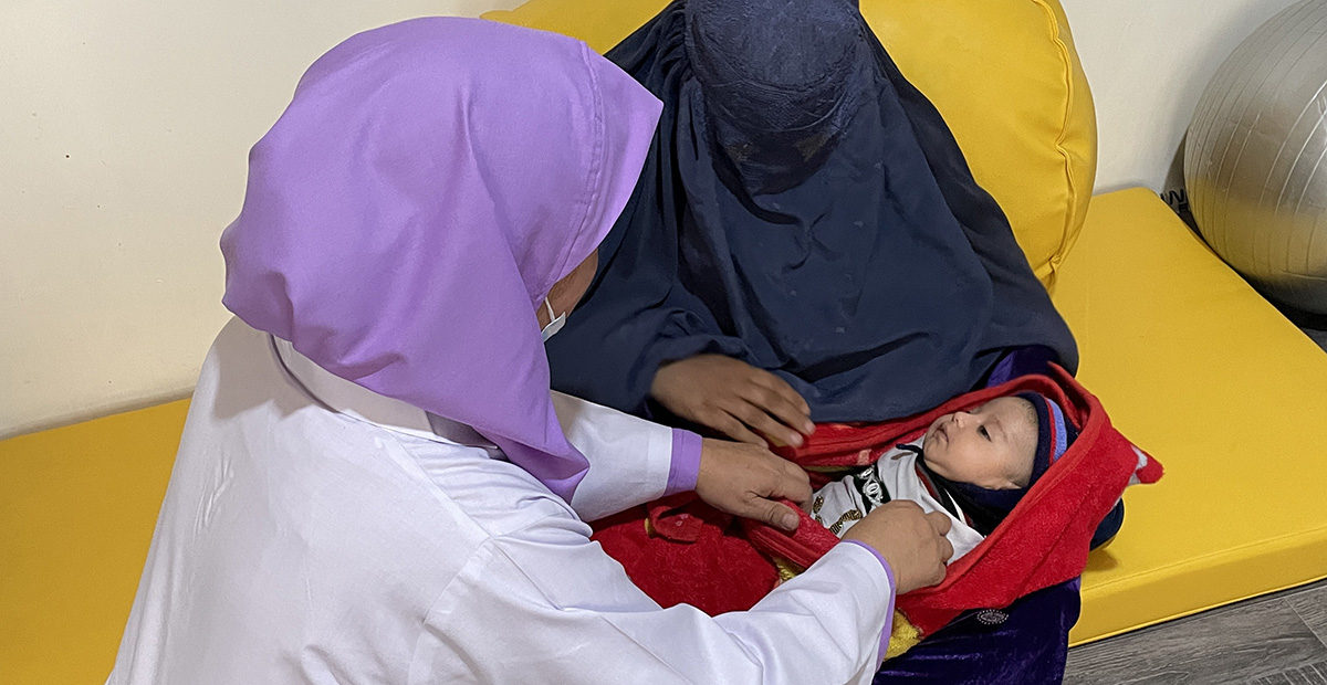 A woman with a purple shawl and a woman covered in a black shawl are caring for a baby wrapped in a red blanket.