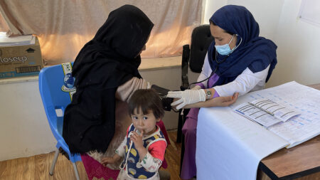 A midwife checks the blood pressure of a patient covered in a shawl. A small child looks into the camera.