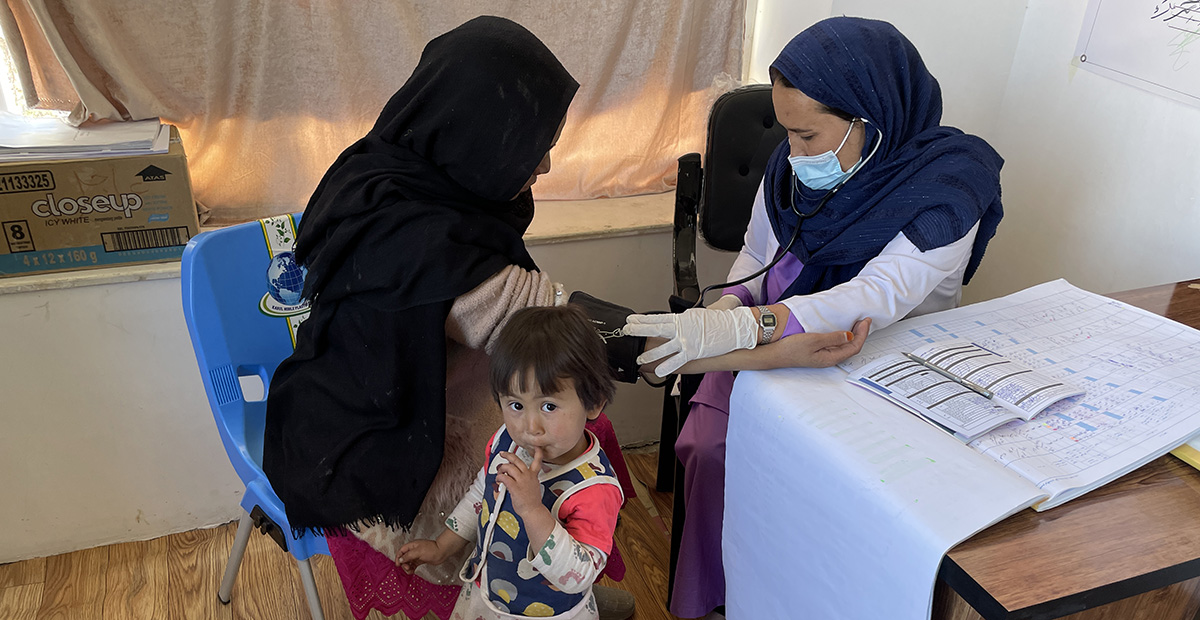 A midwife checks the blood pressure of a patient covered in a shawl. A small child looks into the camera.