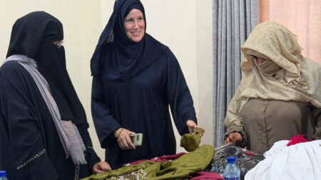 Kajsa Johansson, wearing a shawl and a covering coat, together with two women who have also covered their faces, at a table with hand-sewn garments.