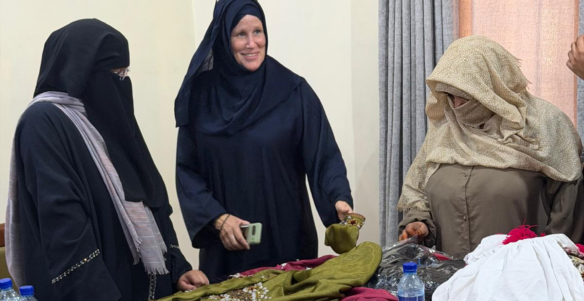 Kajsa Johansson, wearing a shawl and a covering coat, together with two women who have also covered their faces, at a table with hand-sewn garments.