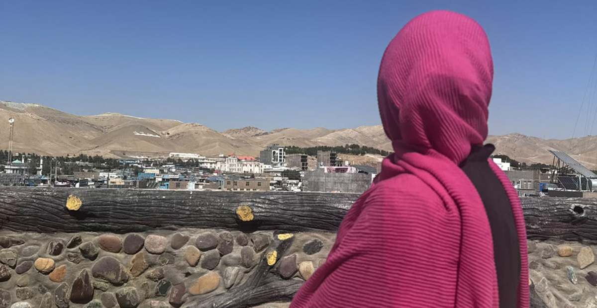 The back of a woman with pink shawl in front of houses and mountains.